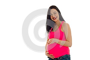 Cheerful young brunette pregnant girl in pink shirt with hand on her belly posing isolated on white background