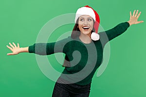 Cheerful young brunette woman wear santa claus hat and wool pullover holding hands wide open over green background.