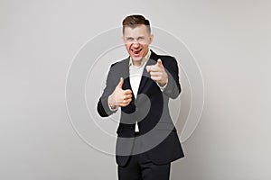Cheerful young business man in classic suit showing thumb up and pointing index fingers on camera isolated on grey
