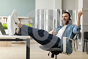 Cheerful young businessman in headphones playing virtual guitar during break in office