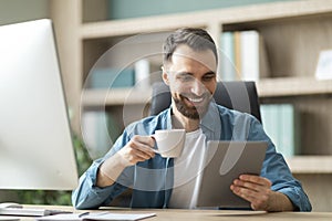 Cheerful young businessman sitting at workdesk, drinking coffee and using digital tablet