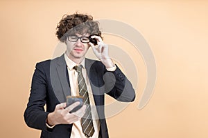 Cheerful young businessman wear spectacles in suit posing surprised and wonder looking at smartphone on orange background. Send or