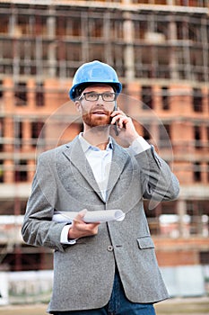 Young civil engineer or businessman standing in front of a construction site and talking on the phone