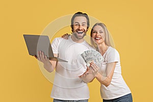Cheerful young caucasian husband and wife in white t-shirts with laptop and many dollars rejoice online victory