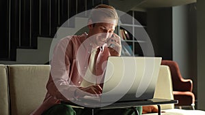 Cheerful young Caucasian man sitting on sofa at desk, working at laptop and talking mobile phone at home. Positive male