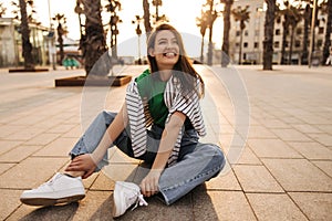 Cheerful young caucasian woman smiles teeth looking to side, sitting in European city on pavement.