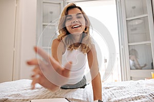 Cheerful young caucasian woman stretched out her hands to camera lying on bed at home.