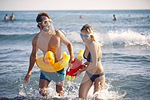 Cheerful young couple on the beach playing with watergun. Man with rubber duck pool float. Summer vacation concept