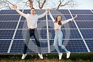 Cheerful young couple jumping together holding hands on background of solar panels under blue sky