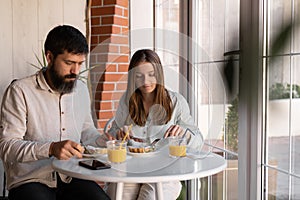 cheerful young couple out for breakfast in a cafe before work, sitting near window and enjoying conversation. woman and