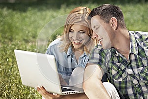 Cheerful young couple sitting on the grass, looking at laptop an