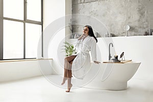 Cheerful young eastern woman in bathrobe sitting on bathtub