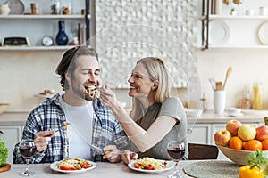 Cheerful young european woman feeds her husband of spaghetti with vegetables or pasta in minimalist kitchen