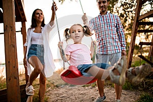 Cheerful young father and mother swing their cute little daughter on swing