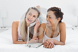 Cheerful young female friends using phone in bed