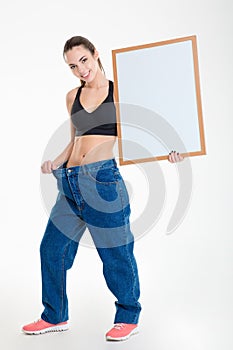 Cheerful young fitness woman in big jeans holding blank board