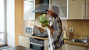 Cheerful young funny woman dancing and singing with lettuce microphone while cooking breakfast