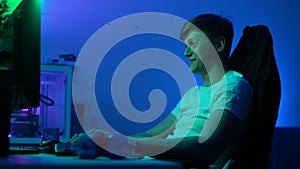 Cheerful young gamer male streaming video games on powerful computer sitting in dark living room with neon lights. Happy