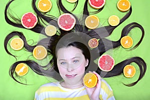 A cheerful young girl lies with citruses in her loose hair, holds a circle of orange in her hands and licks her lips. Top view
