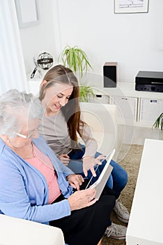 Cheerful young girl sharing time with an old senior woman and teaching internet with computer tablet