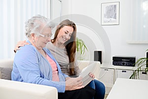 Cheerful young girl sharing time with an old senior woman and teaching internet with a computer tablet
