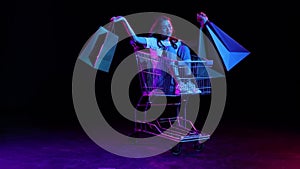 Cheerful young girl sitting in shopping trolley with shopping bags. Concept of business, black friday sales, discount