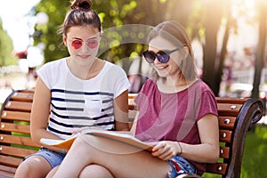 Cheerful young girls spend time together in hot sunny day in the park. Beauties look through youth magazine while sitting on