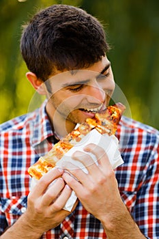 Cheerful young guy is eating pizza on the park