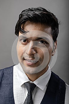 A cheerful, young, handsome Indian male model in formal wear wearing white shirt, black tie, black coat against grey background.
