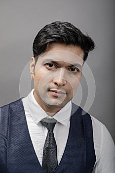 A cheerful, young, handsome Indian male model in formal wear wearing white shirt, black tie, black coat against grey background.