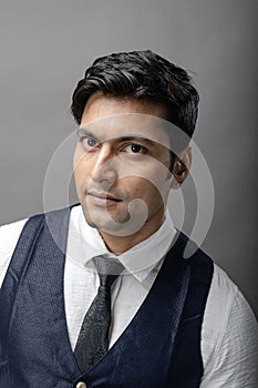 A cheerful, young, handsome Indian male model in formal wear wearing white shirt, black tie, black coat against grey background.