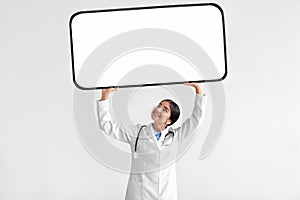 Cheerful young hindu woman doctor in white coat with stethoscope hold huge phone with blank screen above head
