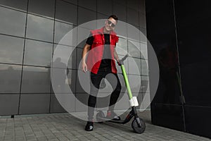 Cheerful young hipster man with a smile in stylish denim red-black clothes in sunglasses stands with a modern electric scooter