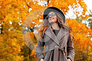 Cheerful young hipster woman in fashionable outerwear in an elegant hat in stylish glasses walks in the park. Happy girl with a