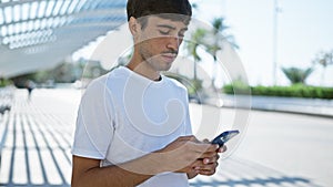 Cheerful young hispanic man joyfully using mobile technology, texting away in the heart of the city park. handsome guy smiling,