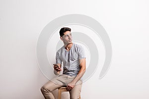 A cheerful young hispanic man with smartphone sitting on a stool in a studio.