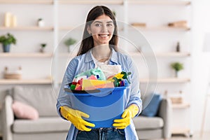 Cheerful young housewife holding bucket with cleaning supplies tools