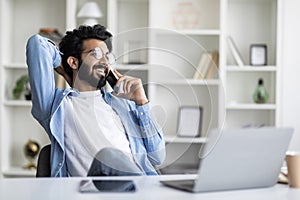 Cheerful Young Indian Guy Talking On Cellphone While Working In Home Office