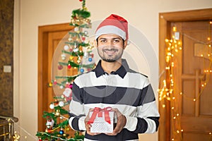 Cheerful Young Indian man holding gift box in hand celebrating christmas alone at home, new year, holiday, party, winter, december