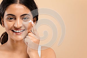 Cheerful young indian woman applying dab of moisturizing cream on her face