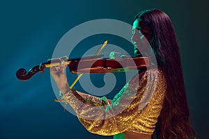 Cheerful young Indian woman, violinist in festive attire performing in neon light against gradient background.