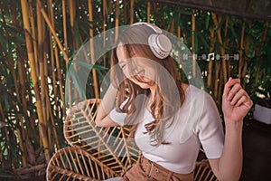 Cheerful young lady in wireless headphones sitting on chair in garden, listening to music, raising her arm, dancing to favorite