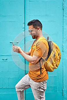 Cheerful young latin guy with his backpack typing on his cell phone walking down the street, wearing orange t-shirt and