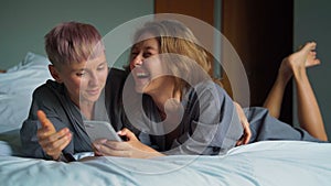 Cheerful young lesbian couple laughing while using their smartphones in the bed