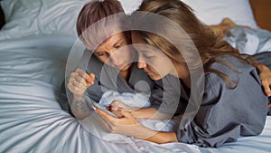 Cheerful young lesbian couple laughing while using their smartphones in the bed