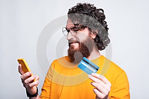 Cheerful young man with beard is shopping online holding his phone and a credit card