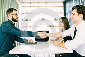 Cheerful young man bonding to his wife while shaking hand to man sitting in front of him at the desk