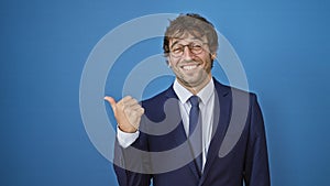 Cheerful young man in business suit, pointing to the side with a thumbs up, smiling, over isolated blue background
