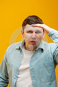 Cheerful young man in casual shirt holding hand at forehead looking far away distance on yellow background in studio. People