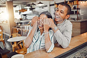 Cheerful young man covering his girlfriends eyes and surprising her while sitting in a cafe. Happy young mixed race